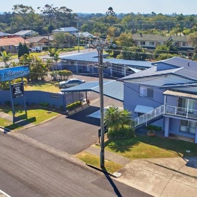An elevated daytime view of the two-story 'Almare Tourist Motel' complex, painted pale blue with a dark blue roof, situated on a street corner in a suburban area.